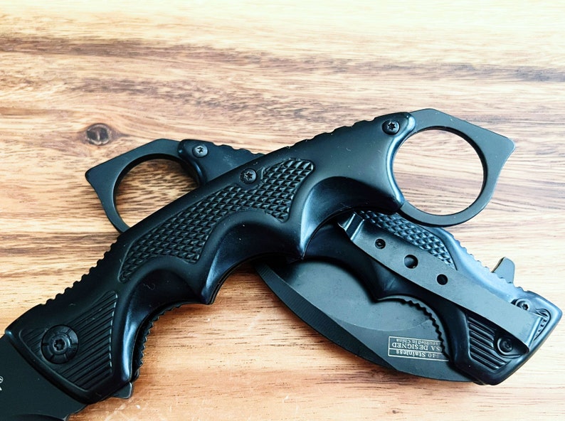 Pu&ograve; includere: Coltello tattico nero con manico ad anello e lama nera. Il coltello ha un'impugnatura testurizzata e un blocco di sicurezza. La lama &egrave; parzialmente aperta e il coltello &egrave; appoggiato su una superficie di legno.