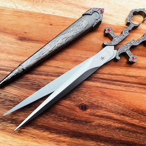 Może przedstawiać: Para ozdobnych srebrnych nożyczek z czerwonymi akcentami. Nożyczki mają ozdobny wzór i pochwę.