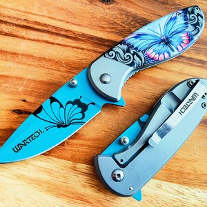 May include: Two blue pocket knives with butterfly designs. The knives have silver blades and handles. The larger knife has the text "WARTECH." on the handle. The smaller knife has the text "WARTECH." on the side of the handle.