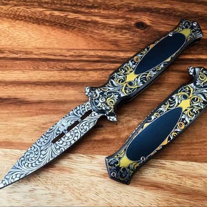 May include: A folding knife with an open blade and a closed handle, both featuring intricate silver and gold scrollwork engravings. The handle has a smooth, dark blue oval inlay centered within the ornate metal pattern and silver screws.