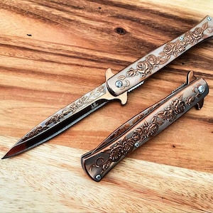 May include: A folding knife with a metallic copper finish and an intricate rose floral engraving on the handle and blade, shown alongside a second identical closed folding knife featuring the same rose pattern and metallic copper-toned finish.
