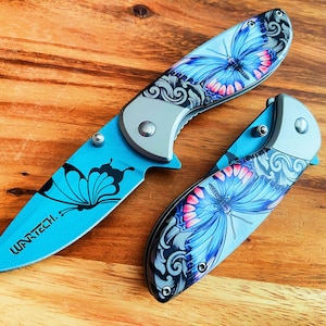 May include: Two blue pocket knives with butterfly designs. The knives have silver handles and black accents. The text "WARTECH." is printed on one of the knives.