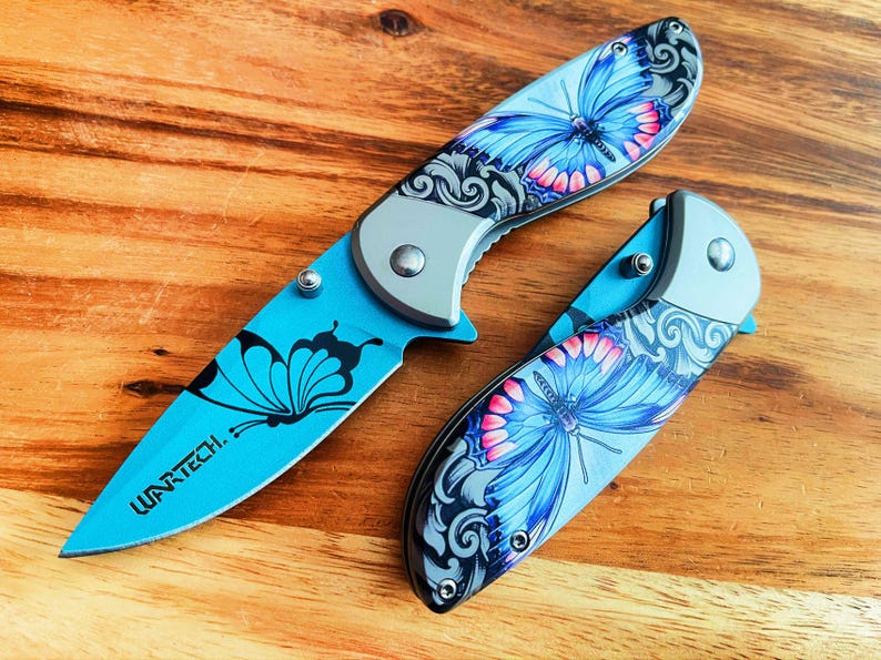 May include: Two blue pocket knives with butterfly designs. The knives have silver handles and black accents. The text "WARTECH." is printed on one of the knives.