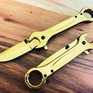 Op de afbeelding: Twee goudkleurige multifunctionele messen met moersleutelhandvatten. De messen hebben een mes en een moersleutel aan elk uiteinde.
