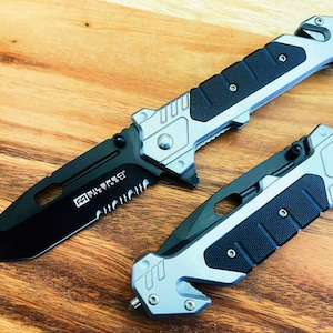 May include: Two silver and black pocket knives with serrated blades and safety features. The knives have a black handle with a textured grip and a silver blade with a black serrated edge. The knives are both closed and have a safety feature on the handle.