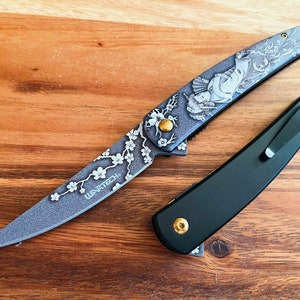 May include: Two pocket knives on a wooden surface. One knife has a gray blade with a floral and geisha design, and the text "WANTECH". The other knife is black with a pocket clip and gold accents.