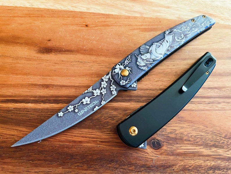 May include: Two pocket knives on a wooden surface. One knife has a gray blade with a floral and geisha design, and the text "WANTECH". The other knife is black with a pocket clip and gold accents.