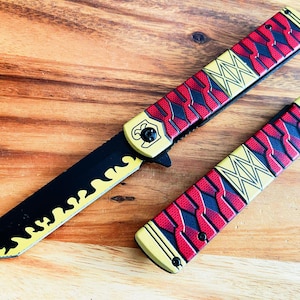 May include: A black and yellow folding knife with a red and yellow patterned handle. The knife is open and the blade is visible. The knife is on a wooden surface.