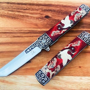 Puede incluir: Una navaja plegable con hoja plateada y mango rojo con diseño de grulla blanca y pino, junto a un mango de navaja cerrado a juego. El refuerzo metálico está decorado con patrones florales blancos y un pequeño logotipo de Falcon.