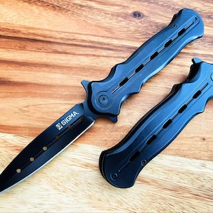 May include: Black folding knife with the word "SIGMA" on the blade. The knife is open, with a pointed blade and a black handle. A second black handle is next to the open knife. The knife is on a wooden surface.