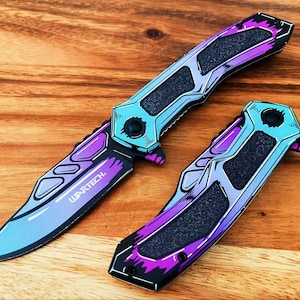Può includere: Due coltelli pieghevoli con una finitura sfumata viola e azzurro e pannelli di presa neri testurizzati. La lama aperta mostra il testo WARTECH. Il design incorpora linee angolari nette e un'estetica metallica in tutto l'oggetto.