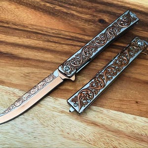 May include: A folding knife with a copper-coloured blade and handle featuring intricate silver-toned scrollwork engravings. The set includes one open knife and one closed knife, both displaying matching ornate floral patterns and metallic finishes.