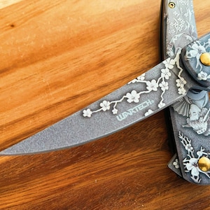 May include: A gray folding knife with intricate floral and geisha designs. The blade features the text "WARTECH" and a cherry blossom branch. The handle has a gold-colored stud. The knife is displayed on a wooden surface.