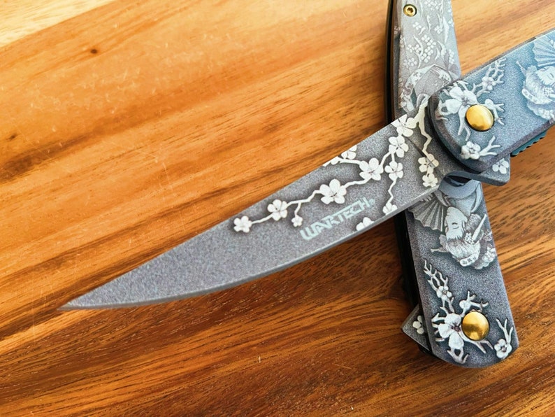 May include: A gray folding knife with intricate floral and geisha designs. The blade features the text "WARTECH" and a cherry blossom branch. The handle has a gold-colored stud. The knife is displayed on a wooden surface.