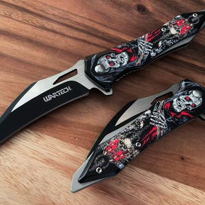 May include: Two folding knives with black blades and silver accents. The handles feature a skull and skeleton design with red and white details. The text "WARTECH" is visible on one blade.