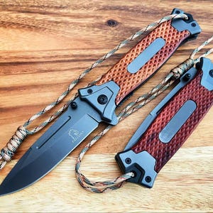 Puede incluir: Dos cuchillos plegables con mangos texturizados marrones y grises y hojas negras se muestran sobre una superficie de madera. Las hojas están parcialmente abiertas, revelando el diseño del cuchillo. Cada cuchillo tiene un cordón adjunto.