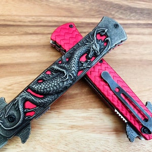 May include: Two red pocket knives with a dragon design on the handle. The knives are open and the blades are visible.