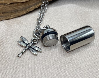 May include: A silver chain necklace with a small silver cylinder pendant that opens. The pendant has a dragonfly charm attached to it. The necklace is lying on a light brown wooden surface.