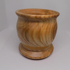 May include: A turned wooden bowl in light brown with a visible wood grain. The bowl has a rounded body, a flared rim, and a pedestal base. The bowl is likely used for decorative purposes, or for holding small items.