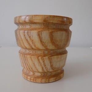May include: A light-coloured wooden vase with a cylindrical shape and a flared base. The vase has a series of horizontal ridges and a smooth, polished finish. The wood grain is visible, showcasing a natural pattern. The vase is suitable for holding flowers or as a decorative item.