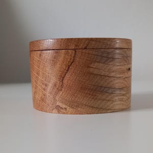 May include: A cylindrical wooden box with a natural light brown finish, featuring a visible wood grain pattern throughout the exterior surface. The container includes a matching circular lid that sits flush with the base, creating a seamless profile.