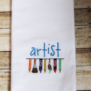 May include: White kitchen towel with blue embroidery that says "artist" and a row of colorful art supplies including paintbrushes, pencils, and crayons.