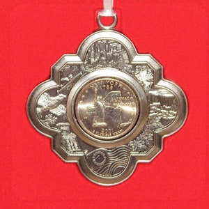 May include: Silver-colored New York state quarter ornament with a white ribbon for hanging. The ornament features the Statue of Liberty, the text "New York 1788", and other state symbols. The ornament is set against a red background.