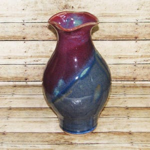 May include: A ceramic vase with a glossy finish featuring a blend of deep magenta and dark blue colors. The vase has a wide, rounded body that tapers toward a narrow neck, ending in a flared, wavy rim. The surface shows smooth color transitions.