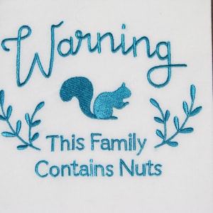 May include: A white kitchen towel with teal blue embroidery. The embroidery says "Warning This Family Contains Nuts" and features a teal blue squirrel.