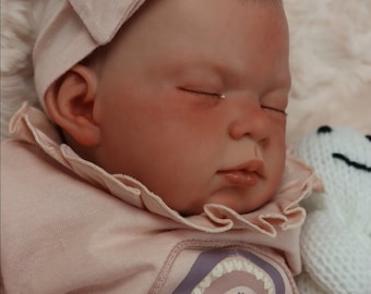 May include: A sleeping baby doll wearing a pink ruffled onesie with a rainbow design and a pink bow headband.