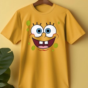 Op de afbeelding: Mosterdgeel T-shirt met een groot, lachend cartoongezicht. Het gezicht heeft blauwe ogen, witte tanden en groene stippen. Het shirt met korte mouwen hangt aan een houten hanger.