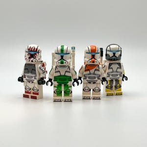 Puede incluir: Cuatro figuras de juguete de soldados clon en armadura blanca con detalles de color. Cada figura tiene un diseño de casco único con detalles rojos, verdes, naranjas y grises. Las figuras están sobre una superficie blanca.