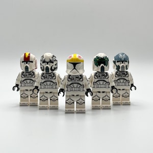 Peut inclure: Cinq figurines Lego blanches de soldats clones avec des casques détaillés. Chaque figurine a un design de casque différent, avec des couleurs comprenant des accents rouges, noirs, jaunes, verts et bleus. Les figurines sont alignées.