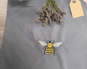 May include: A gray tote bag with a bee embroidered on the front. The bee is yellow and black with white wings. The bag has two handles and a brown tag attached to one of the handles.