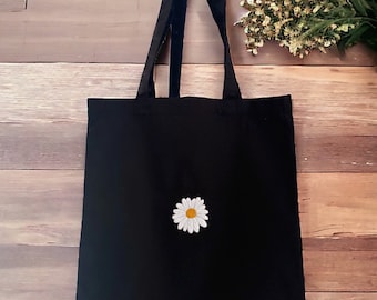 May include: A black tote bag featuring a single embroidered daisy flower centered on the front. The flower has white petals and a yellow center. The bag includes two black fabric shoulder straps attached to the top edge of the rectangular body.