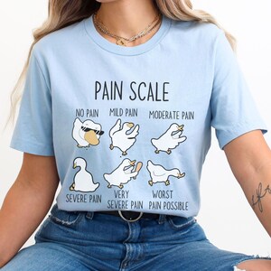 Pu&ograve; includere: Una maglietta blu chiaro con una grafica di una scala del dolore che utilizza anatre dei cartoni animati. Il testo sulla maglietta recita "Pain Scale", "No Pain", "Mild Pain", "Moderate Pain", "Severe Pain", "Very Severe Pain", e "Worst Pain Possible".