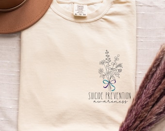 May include: Cream-colored t-shirt with a floral design and the words "Suicide Prevention awareness." The design includes a bouquet of flowers with a teal and purple ribbon. A brown hat is visible in the upper left corner.