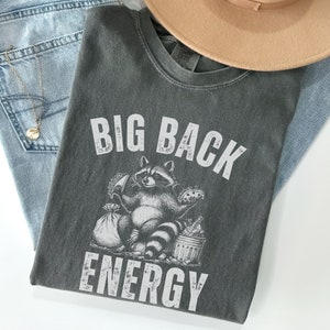 May include: Dark gray t-shirt with the text "BIG BACK ENERGY" and a raccoon graphic. The raccoon is holding food and standing next to a trash can. The shirt is folded on top of blue jeans and a tan hat.