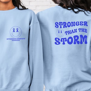 May include: Light blue sweatshirt with a blue smiley face with two blue ribbons for eyes and the text "Osteogenesis Imperfecta Awareness" on the front. The back of the sweatshirt has the text "Stronger Than The Storm" in blue with a blue smiley face with two blue ribbons for eyes.