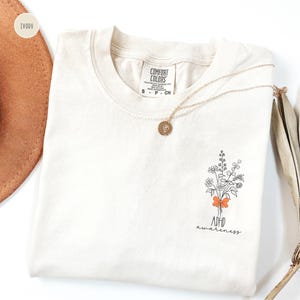 May include: Ivory-colored t-shirt with a floral bouquet graphic and the words "ADHD awareness." A gold necklace with a round pendant rests on the shirt. The shirt is folded, and a brown hat is partially visible in the background.