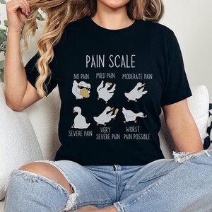Pu&ograve; includere: Una maglietta nera con una grafica bianca di una scala del dolore che utilizza oche. La scala va da "Nessun dolore" a "Peggior dolore possibile".