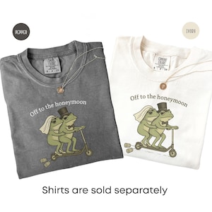 May include: Two short-sleeve t-shirts in pepper gray and ivory, each featuring a graphic of two frogs riding a scooter with the text Off to the honeymoon. The frog bride wears a veil and necklace, while the frog groom wears a top hat and bow tie.