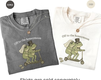 May include: Two short-sleeve t-shirts in pepper gray and ivory, each featuring a graphic of two frogs riding a scooter with the text Off to the honeymoon. The frog bride wears a veil and necklace, while the frog groom wears a top hat and bow tie.