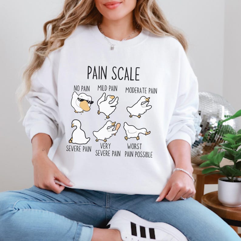 May include: White sweatshirt with a graphic of a pain scale using cartoon geese. The text reads "Pain Scale" with different levels of pain from "No Pain" to "Worst Pain Possible".