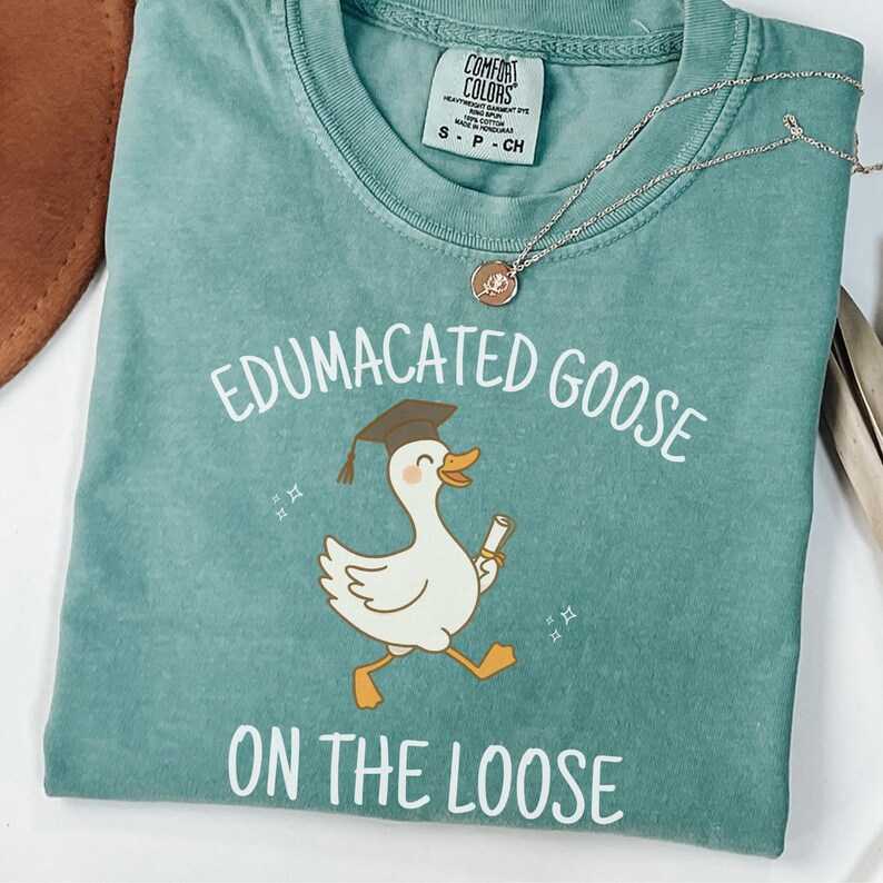 K&ouml;nnte beinhalten: Ein blaugr&uuml;nes T-Shirt mit einer wei&szlig;en Grafik einer Gans, die einen Abschluss-Hut tr&auml;gt und ein Diplom h&auml;lt. Der Text "Edumacated Goose on the Loose" ist auf das Shirt gedruckt.