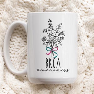 May include: White ceramic coffee mug with a black line drawing of a bouquet of flowers tied with pink and teal ribbons. The text "BRCA awareness" is printed below the flowers.