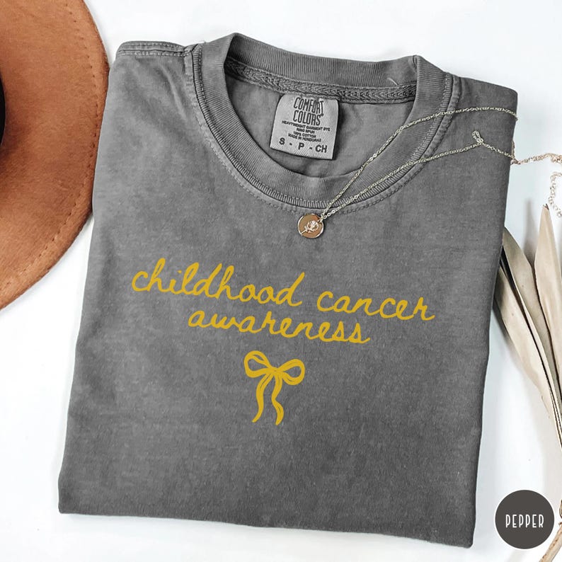 May include: A charcoal grey t-shirt featuring the yellow text "childhood cancer awareness" in a cursive script font above a yellow ribbon graphic. The shirt has a Comfort Colors brand label at the inner neckline and a silver-toned pendant necklace.