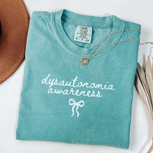 May include: Teal blue t-shirt with the text "dysautonomia awareness" in white script with a white bow underneath.