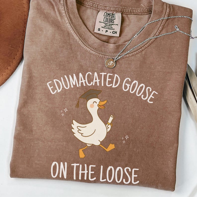 K&ouml;nnte beinhalten: Ein braunes T-Shirt mit dem Text "Edumacated Goose on the Loose" und einer Cartoon-Illustration einer Gans, die einen Abschluss-Hut tr&auml;gt und ein Diplom h&auml;lt.