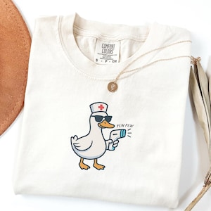 May include: Cream-colored t-shirt featuring a cartoon duck wearing sunglasses and a nurse's hat, holding a thermometer gun. The text "PEW PEW" is printed next to the gun. A gold necklace and the Comfort Colors tag are also visible.